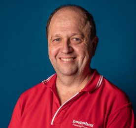 A man smiling in a red collared shirt standing before a blue background.