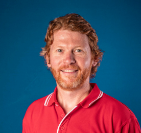 Smiling man with curly red hair and beard wearing a red polo shirt, posed against a blue background.