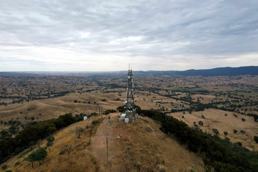 Aerial view of a telecommunications tower and equipment shelters on a rural hilltop surrounded by farmland and rolling hills.