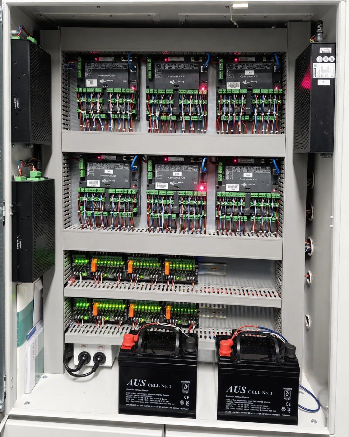 Open industrial electrical control cabinet showing multiple black PLC modules, green terminal blocks, wiring, and two black batteries labeled AUS CELL