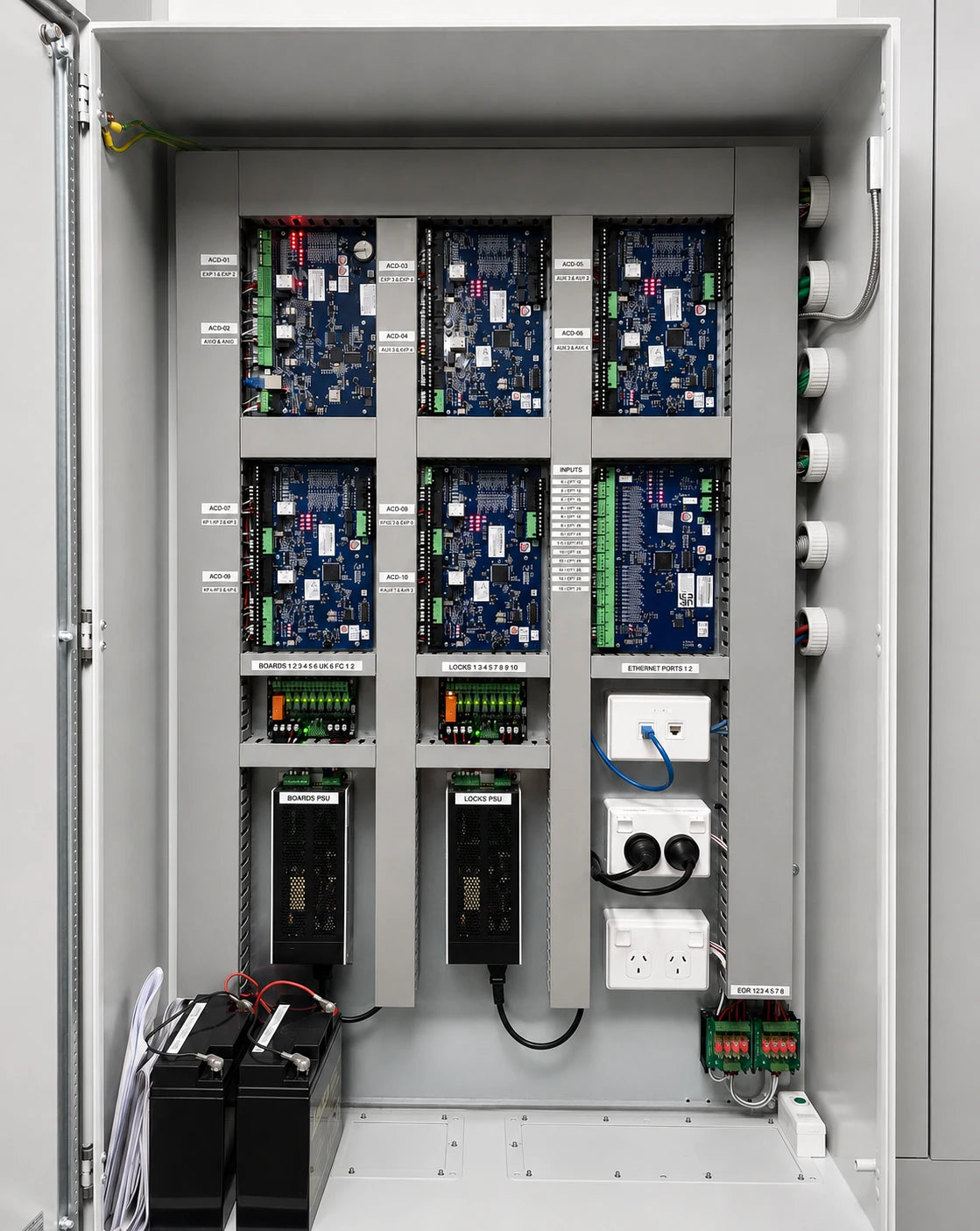 Open industrial electrical control cabinet showing six blue circuit boards arranged in a 3x2 grid, with terminal blocks, power outlets, cables, and two batteries inside a gray metal enclosure.